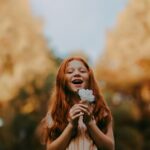 Smiling redheaded girl enjoys nature holding a flower outdoors during the day.