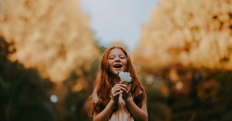 Smiling redheaded girl enjoys nature holding a flower outdoors during the day.